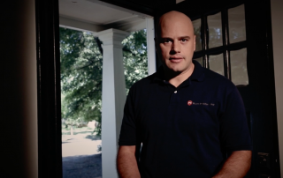 Inspector Adam wearing a blue polo standing near the open front door of a home with front yard in the background