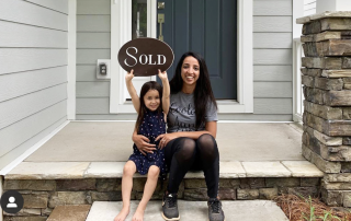 Lara Bucci wearing a gray shirt and black leggings sitting on the front porch of a gray siding home with a blue front door